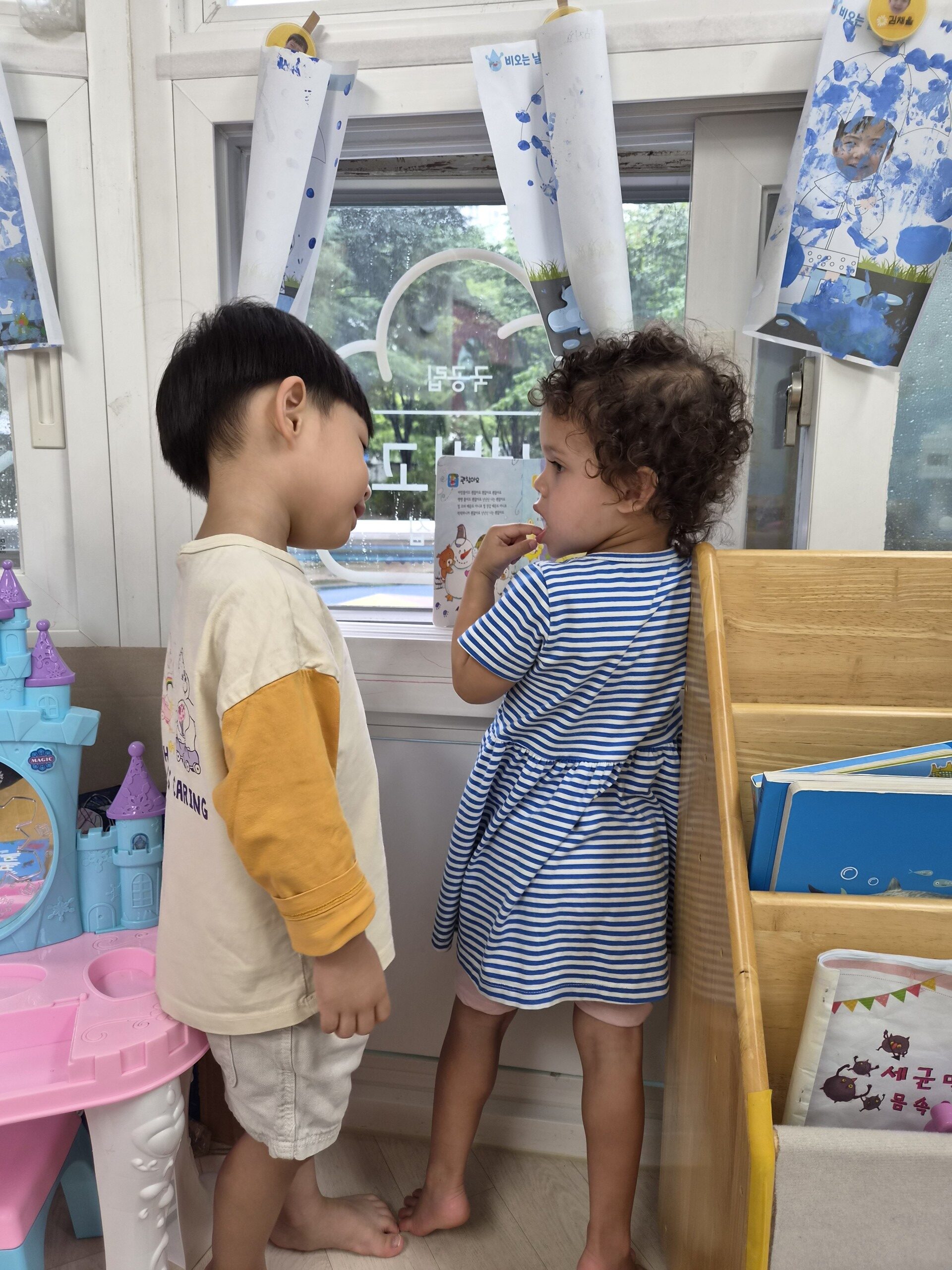 어린이집 창가에 서서 곱슬머리 친구와 마주 보며 다정하게 이야기하는 지호의 모습. 창밖으로 초록색 나무들이 보인다.