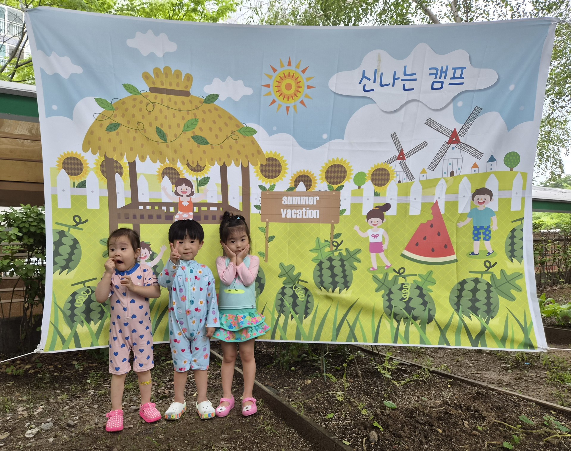 세 명의 어린아이들이 야외에서 '신나는 캠프' 현수막 앞에 나란히 서서 카메라를 보고 있다. 지호는 가운데에서 연한 하늘색 수영복을 입고 손가락으로 브이 포즈를 하며 환하게 웃고 있고, 양옆의 친구들도 각각 귀여운 포즈를 취하고 있다.