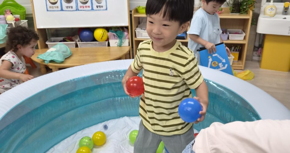 노란색 줄무늬 티셔츠를 입은 지호가 파란색 볼풀장 안에서 빨간 공과 파란 공을 양손에 들고 미소 지으며 서 있다. 주변에는 다양한 색깔의 공들이 흩어져 있고, 다른 아이들도 함께 놀고 있다.