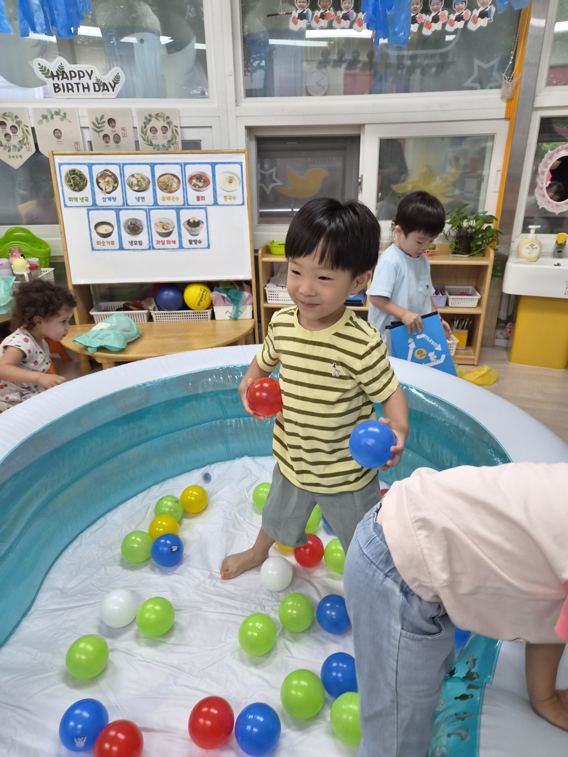 노란색 줄무늬 티셔츠를 입은 지호가 파란색 볼풀장 안에서 빨간 공과 파란 공을 양손에 들고 미소 지으며 서 있다. 주변에는 다양한 색깔의 공들이 흩어져 있고, 다른 아이들도 함께 놀고 있다.