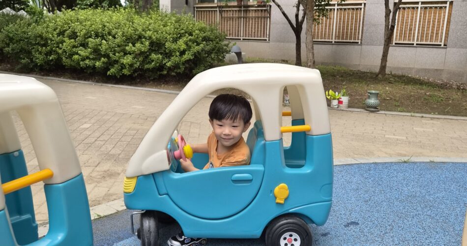 푸른색 놀이터 바닥 위, 파란색 미니카에 앉아 정면을 보며 환하게 웃는 지호의 모습. 주황색 반팔 티셔츠를 입고 있으며, 뒤로는 나무와 아파트 건물이 보인다.