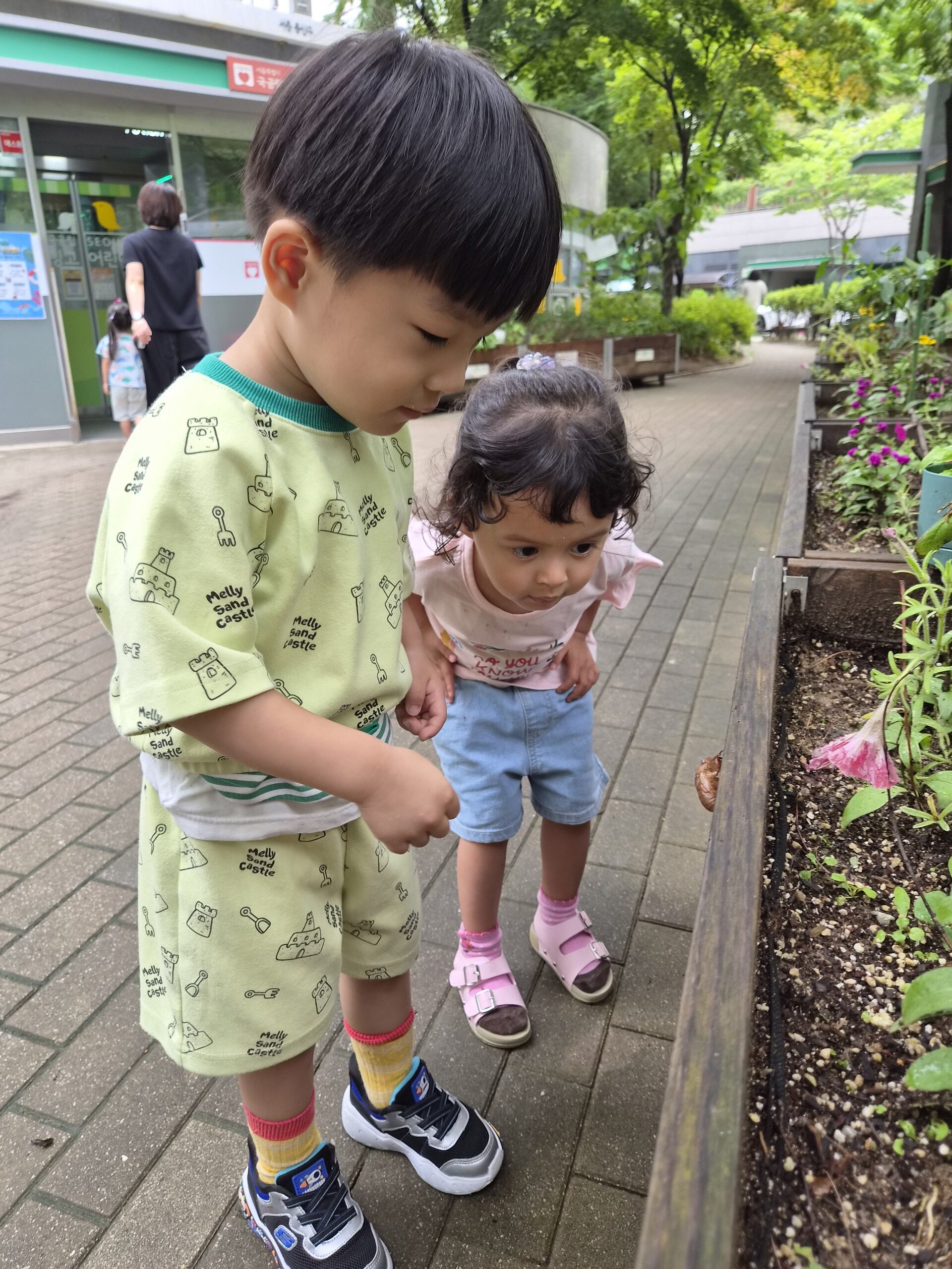 지호와 여자아이가 나란히 서서 나무 화단 옆에 있는 작은 달팽이를 발견하고 신기한 듯 쪼그리고 앉아 집중해서 바라보고 있다. 지호는 연두색 옷을, 친구는 분홍색 티셔츠와 청바지를 입고 있다.