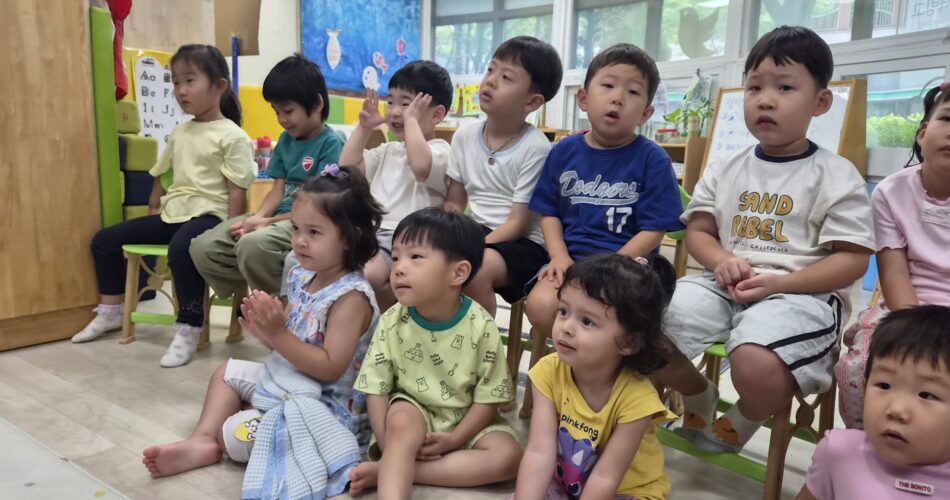 어린이집 교실에서 여러 명의 아이들이 바닥과 작은 의자에 앉아 한 방향을 바라보고 있는 모습입니다. 아이들은 집중하며 무언가를 경청하고 있는 듯하며, 지호도 그 중 한 명으로 친구들과 함께 있습니다.