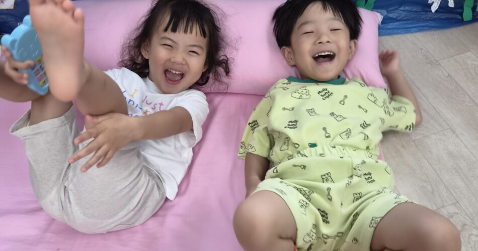 분홍색 매트 위에서 지호와 한 여자아이가 서로 마주 보며 배시시 웃고 있다. 지호는 연두색 잠옷을 입고 노란색 줄무늬 양말을 신었고, 여자아이는 하얀색 티셔츠를 입고 다리를 들고 있다. 뒤편에는 바닷속 풍경 그림이 걸려 있다.