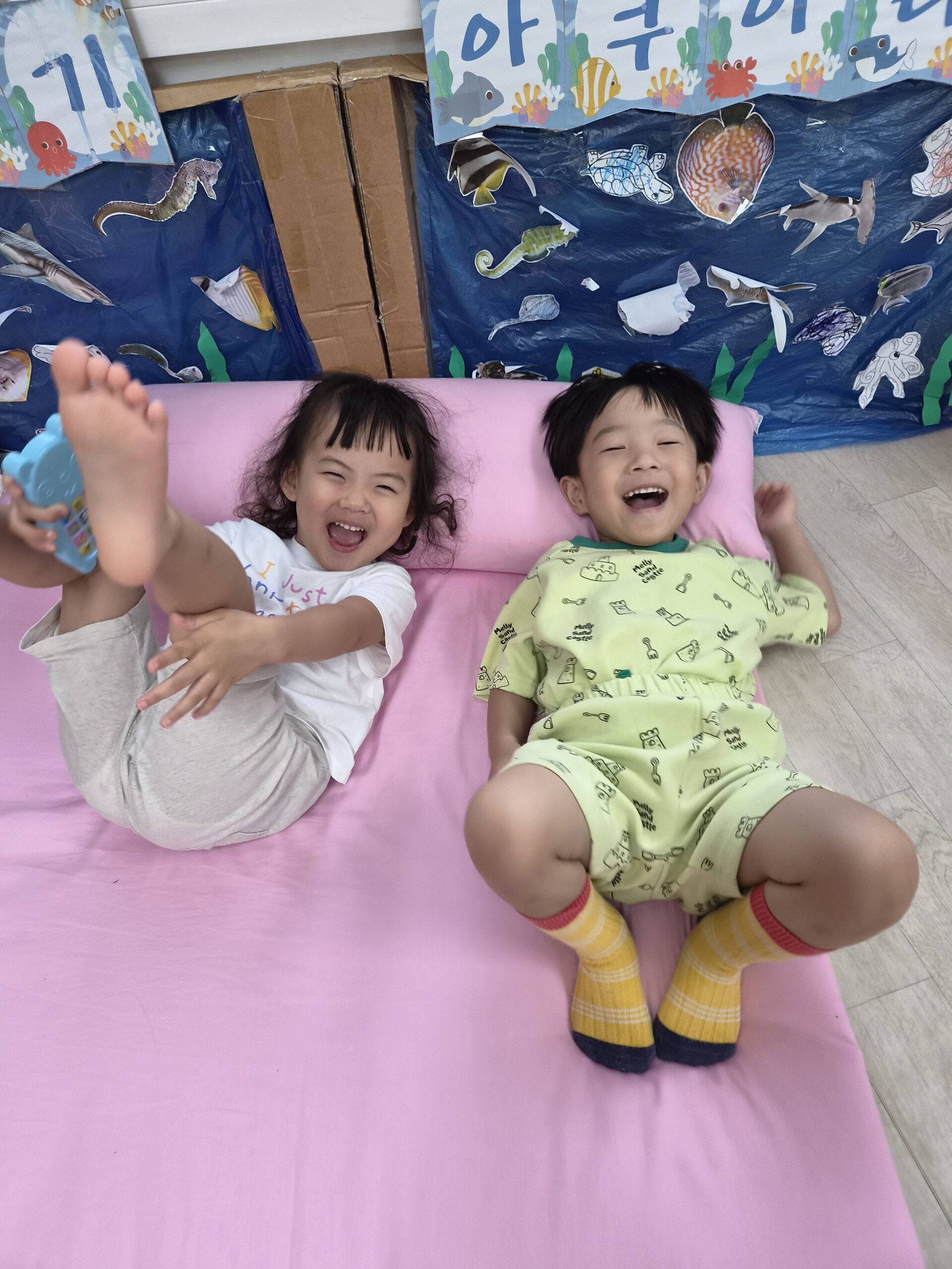 분홍색 매트 위에서 지호와 한 여자아이가 서로 마주 보며 배시시 웃고 있다. 지호는 연두색 잠옷을 입고 노란색 줄무늬 양말을 신었고, 여자아이는 하얀색 티셔츠를 입고 다리를 들고 있다. 뒤편에는 바닷속 풍경 그림이 걸려 있다.