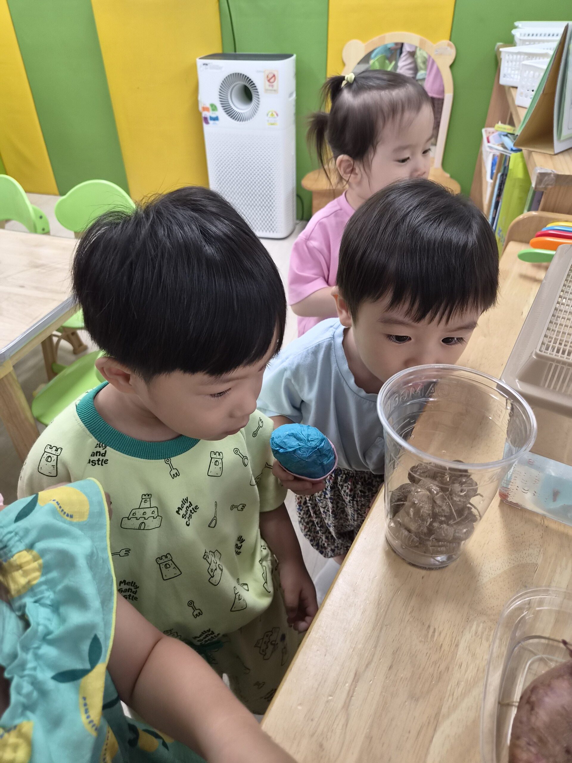 어린이집 교실에서 지호와 친구가 나무 탁자 위에 놓인 투명 컵 속의 말린 나뭇잎이나 알갱이들을 호기심 어린 눈으로 자세히 들여다보고 있다. 지호는 파란색 조형물을 들고 있다.