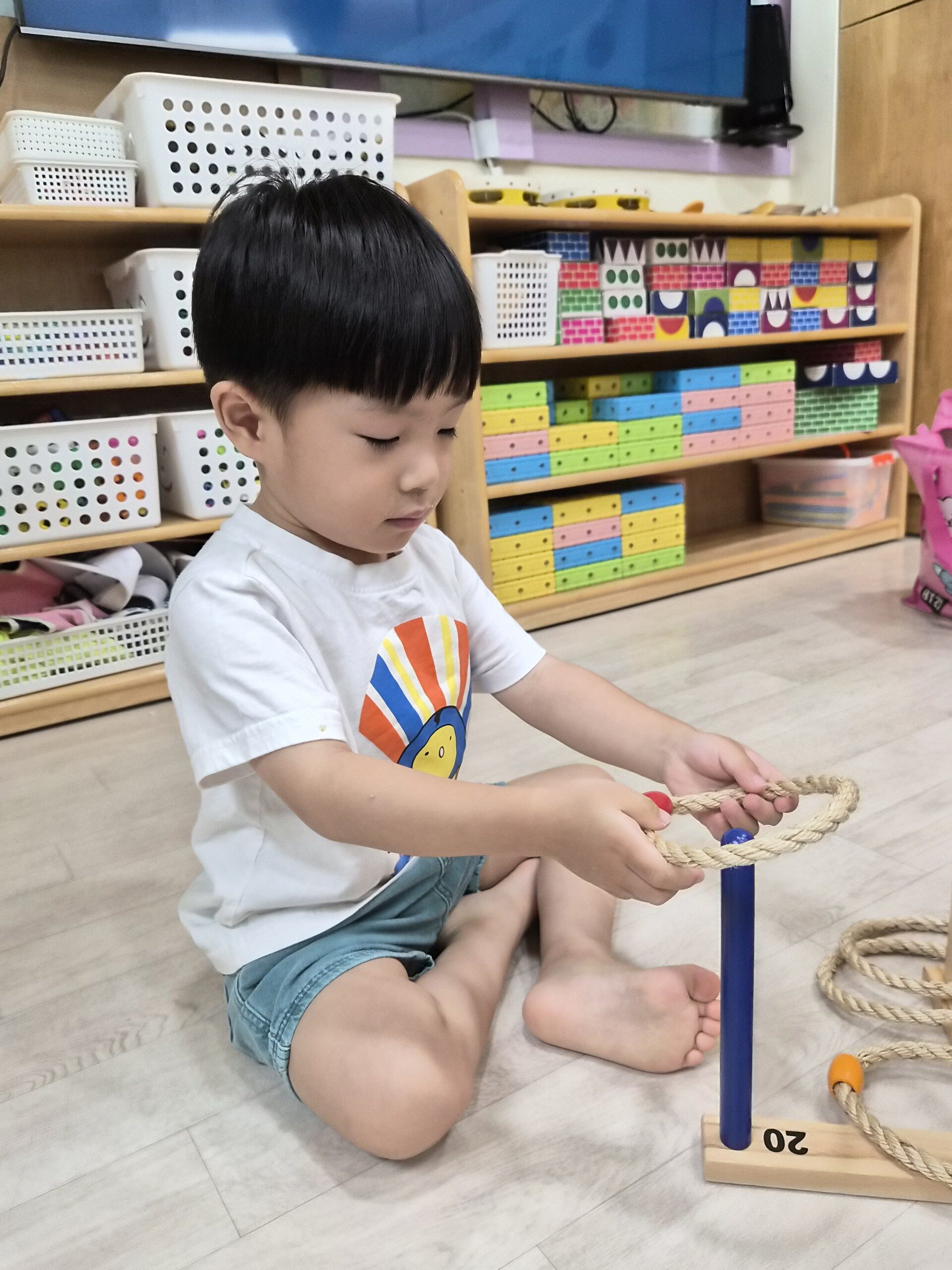 실내 마루 바닥에 앉아있는 지호가 하얀색 티셔츠를 입고 고리 던지기 놀이에 집중하고 있다. 양손으로 노란색 고리를 잡고 파란색 기둥에 끼우려 하고 있으며, 뒤쪽으로는 다양한 장난감이 정리된 선반이 보인다.