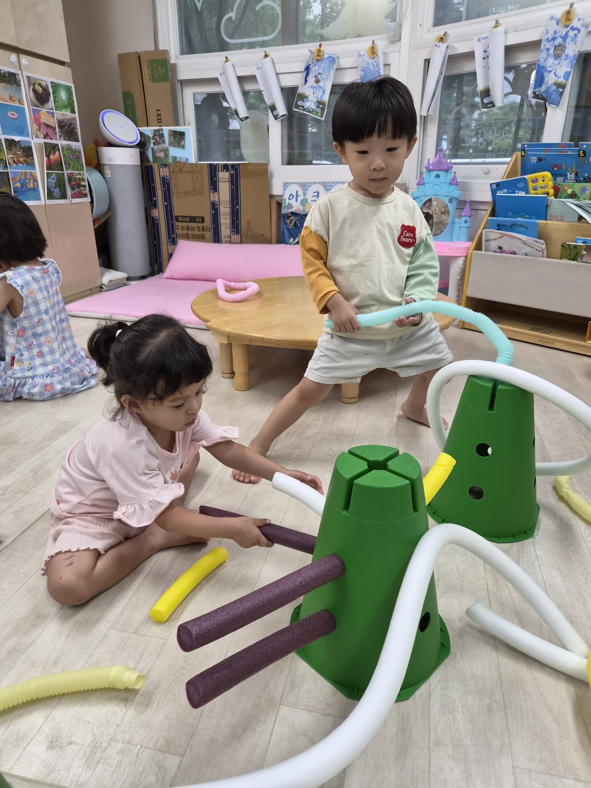 어린이집 놀이방에서 지호가 서서 파란색 파이프를 들고 있고, 옆에 앉은 여자아이는 다른 파이프와 녹색 블록으로 놀이에 집중하고 있다.