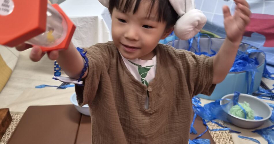 베이지색 옷과 하얀 털모자를 쓴 지호가 밝게 웃으며 빨간색 모래시계 장난감을 앞으로 내밀고 있다. 배경에는 파란색 '목욕탕' 장식이 보인다.