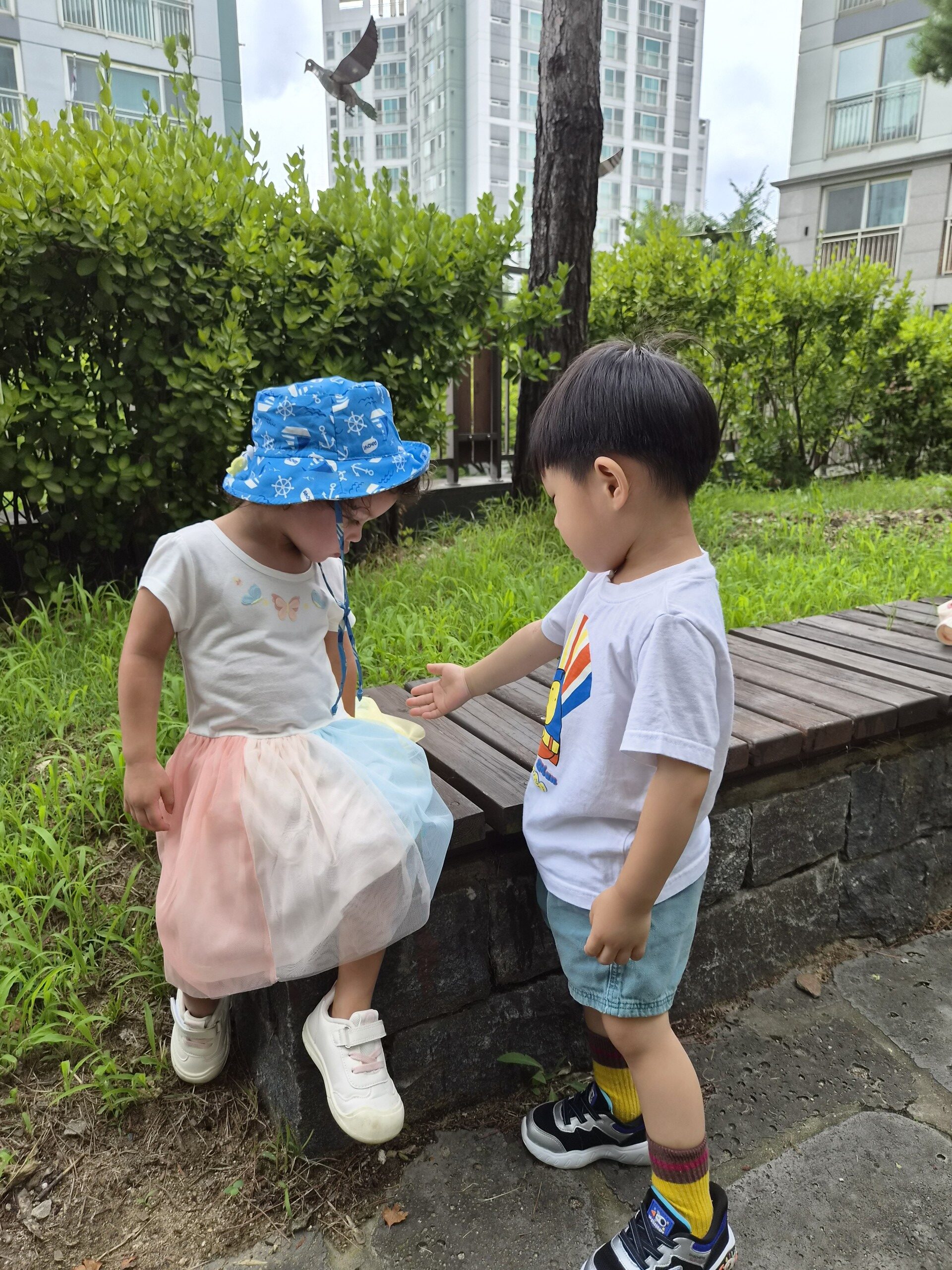야외 벤치에 앉아있는 파란색 모자와 무지개 치마를 입은 여자아이에게 지호가 하얀색 티셔츠와 파란색 반바지를 입고 다가가 손을 내밀며 이야기하고 있다. 뒤편으로는 아파트와 나무들이 보인다.