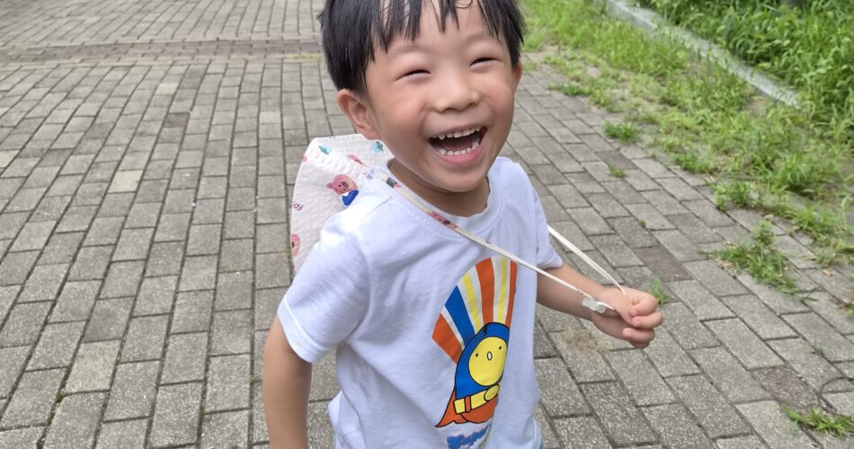 야외 포장된 길 위에서 활짝 웃으며 카메라를 향해 달려오는 지호의 모습. 흰색 반팔 티셔츠와 파란색 반바지, 알록달록한 양말과 운동화를 신고 있으며, 등 뒤에는 작은 가방이 메어져 있다. 배경에는 푸른 나무와 멀리 다른 아이의 모습이 보인다.