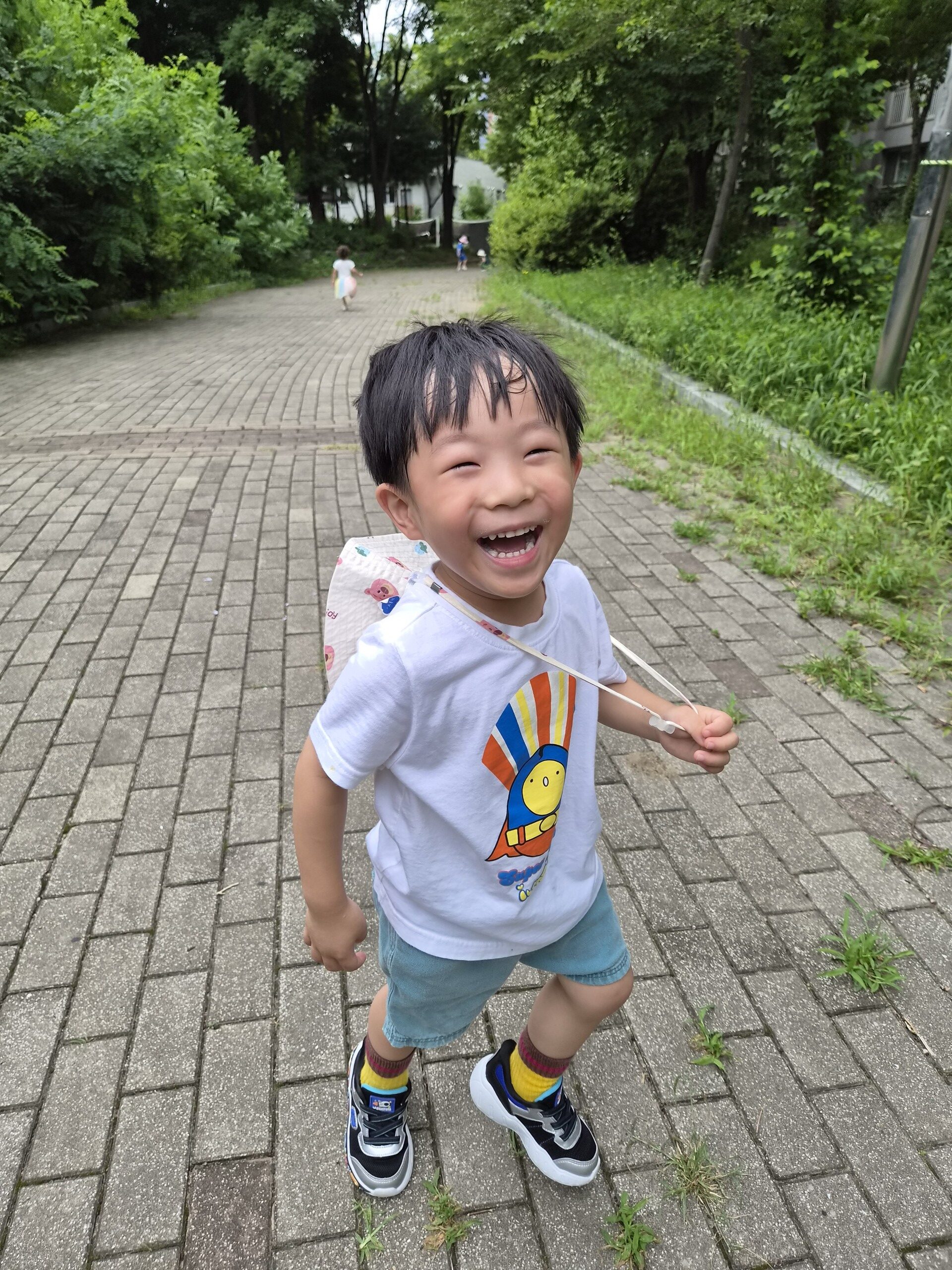야외 포장된 길 위에서 활짝 웃으며 카메라를 향해 달려오는 지호의 모습. 흰색 반팔 티셔츠와 파란색 반바지, 알록달록한 양말과 운동화를 신고 있으며, 등 뒤에는 작은 가방이 메어져 있다. 배경에는 푸른 나무와 멀리 다른 아이의 모습이 보인다.
