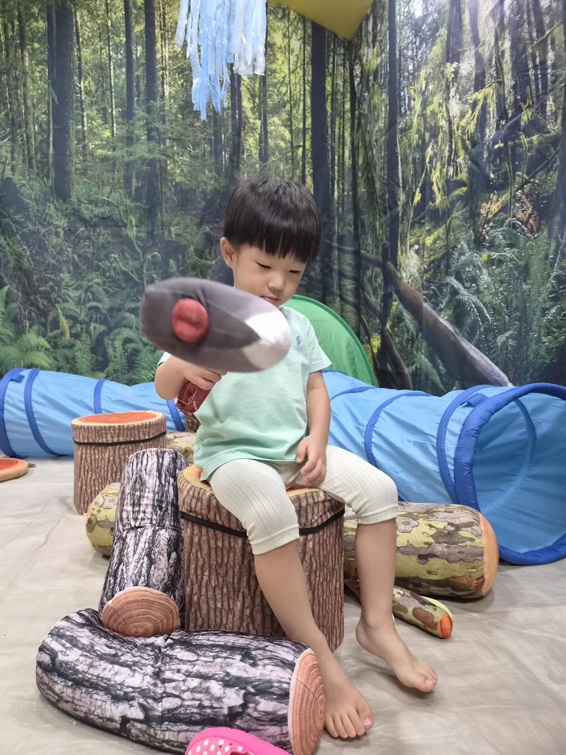 숲 배경 앞에서 지호가 통나무 모양의 쿠션 의자에 앉아 검은색과 빨간색 장난감 도끼를 들고 아래를 내려다보고 있습니다. 파란색 터널이 뒤쪽에 보입니다.
