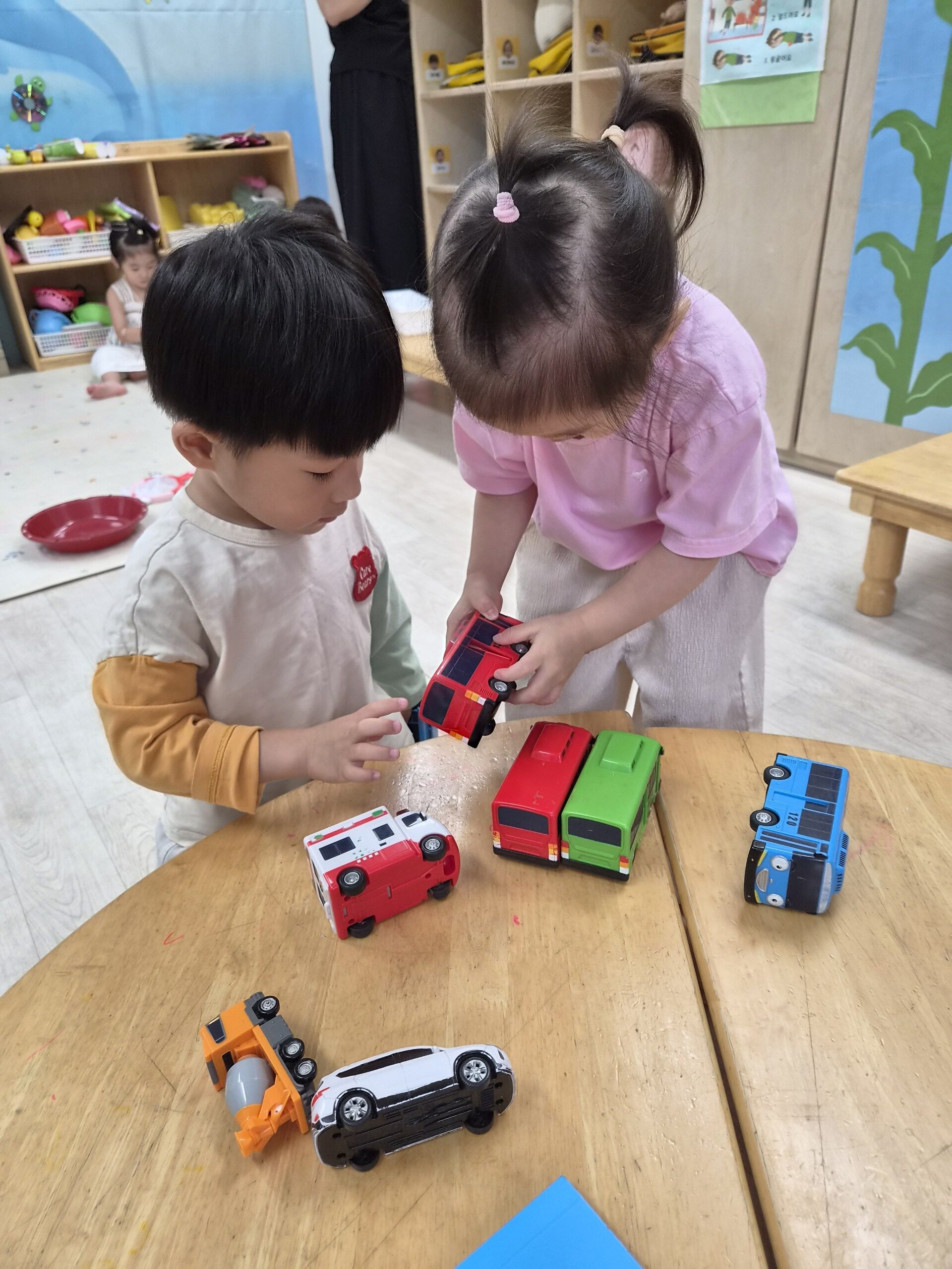 지호와 여자아이가 나무 테이블에 앉아 빨간색, 초록색, 파란색 등 다양한 색상의 변신 로봇 자동차 장난감들을 가지고 놀고 있습니다. 지호는 왼쪽 손으로 빨간색 자동차를 만지고 있습니다.