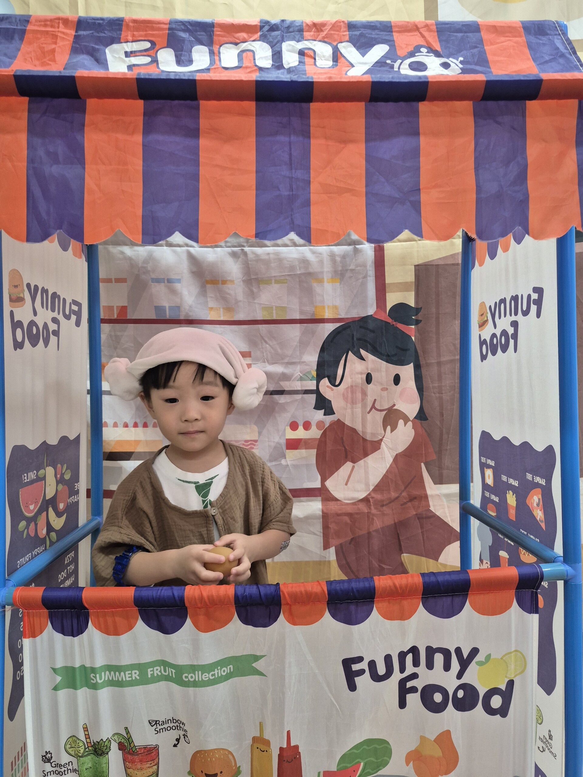 주황색과 보라색 줄무늬 지붕을 가진 'Funny Food'라고 쓰인 플레이 텐트 안에서 지호가 분홍색 귀 달린 모자를 쓰고 앉아 있다. 지호는 갈색 상의를 입고 손에 작은 물건을 들고 있으며, 텐트 벽에는 음식 그림들이 그려져 있다.