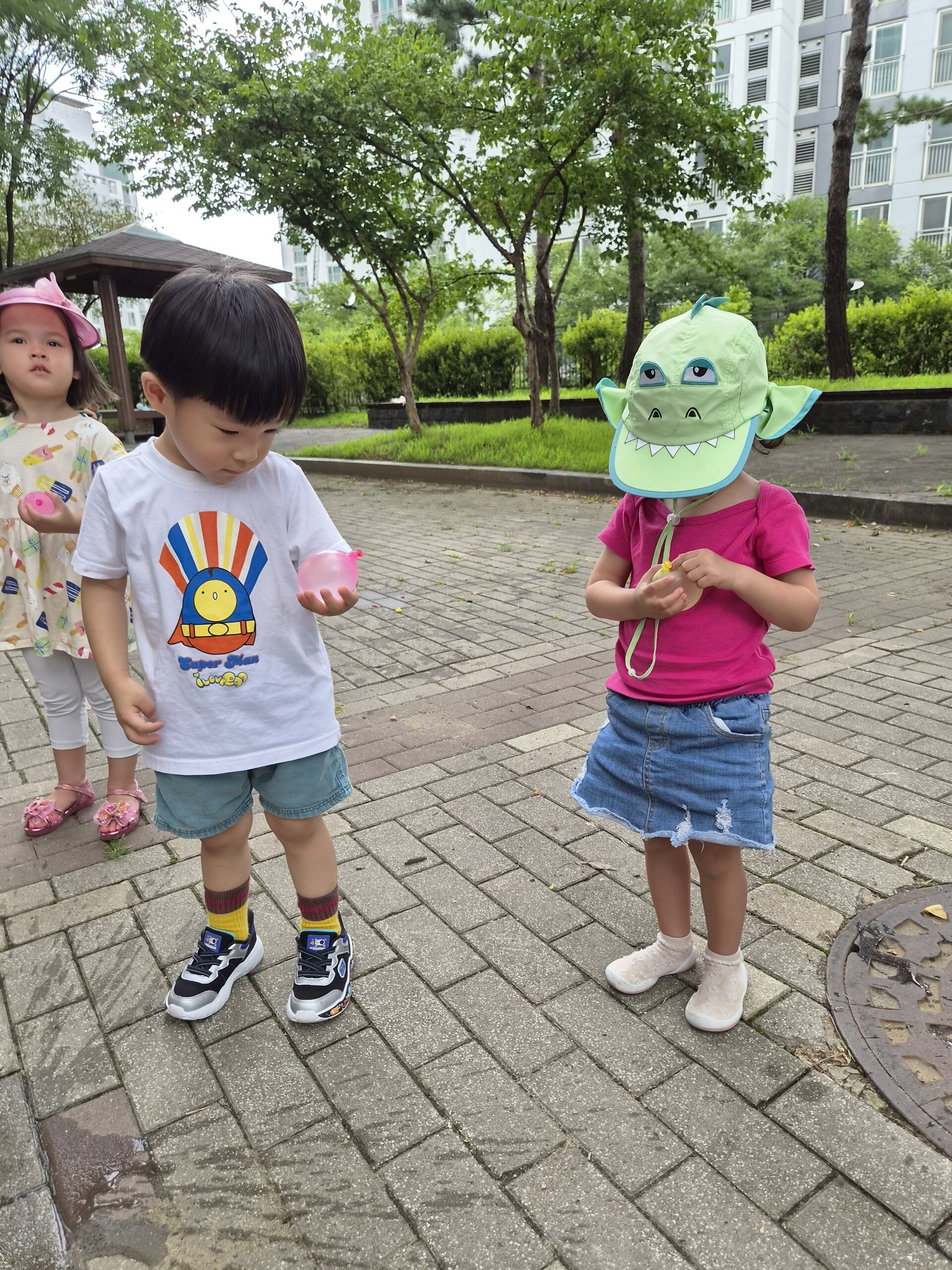 하얀색 기차 그림 티셔츠를 입은 지호와 두 명의 여자아이가 야외 공원 보도블록 위에서 나란히 서 있다. 한 친구는 공룡 모자를 쓰고 있다.