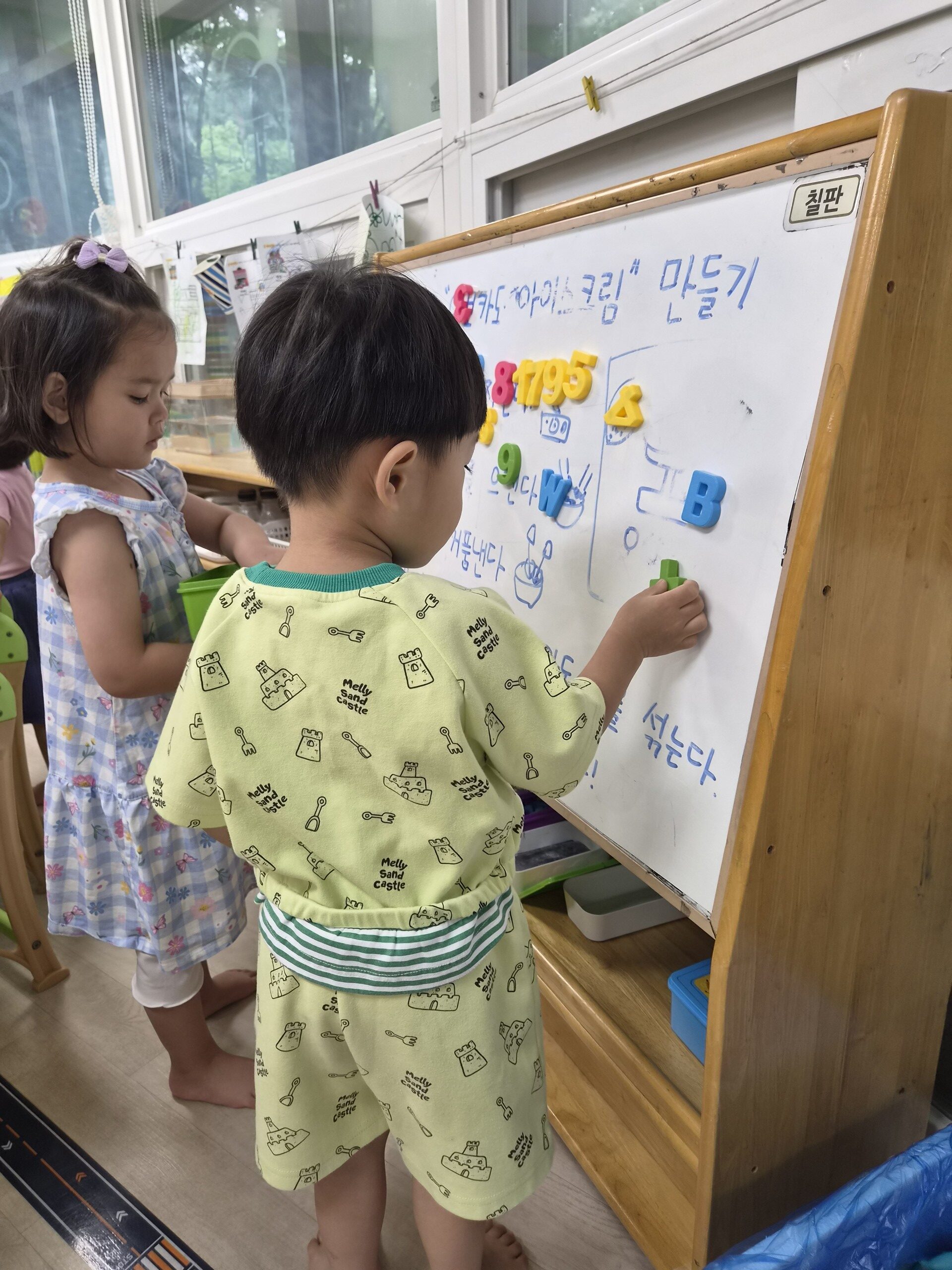지호가 어린이집 교실에서 하얀색 칠판 앞에 서서 자석 글자 'L'을 칠판에 붙이고 있다. 옆에는 다른 친구가 함께 서서 지켜보고 있으며, 칠판에는 여러 자석 글자와 그림이 붙어있다.
