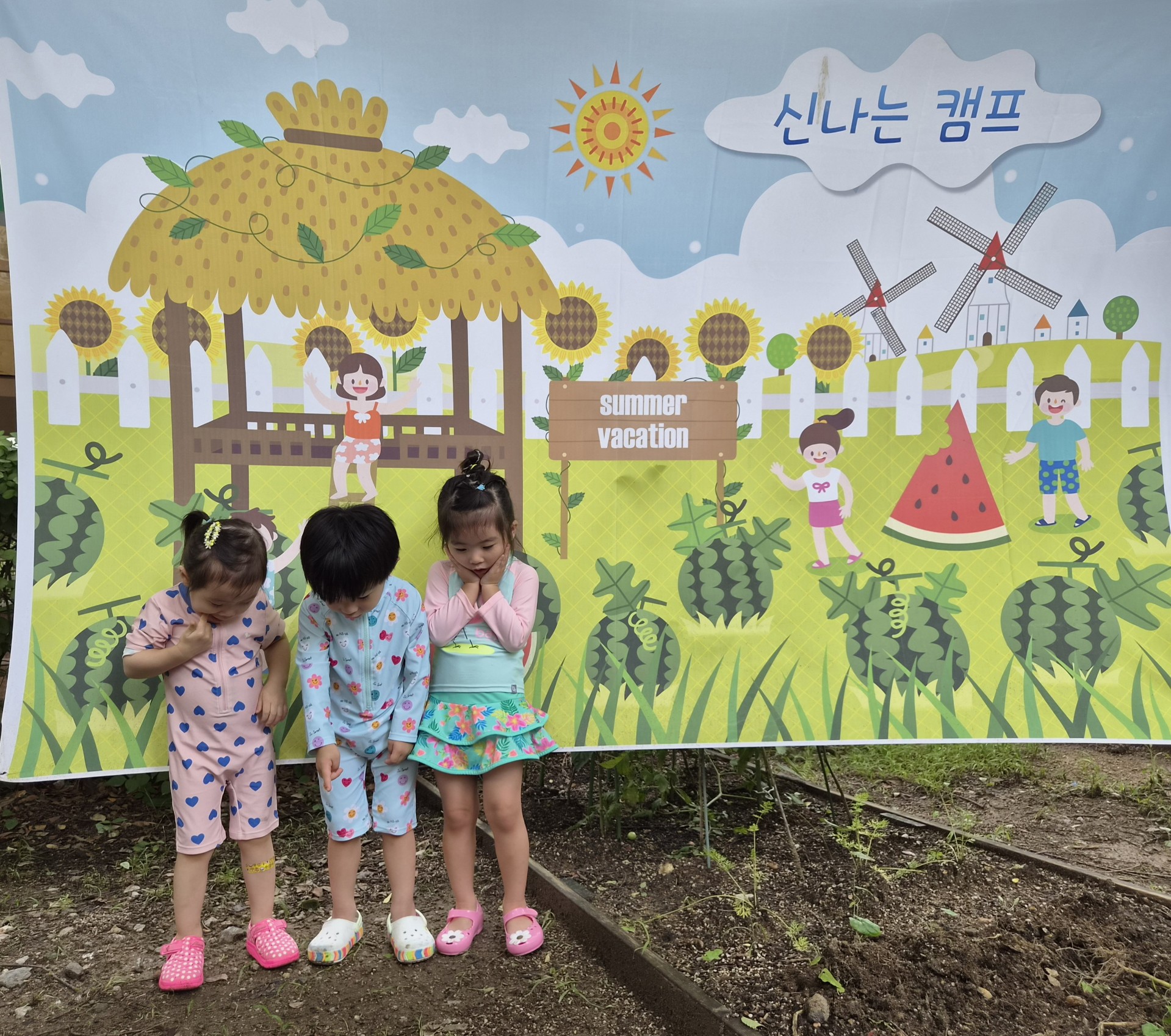 세 명의 어린아이들이 야외에서 '신나는 캠프'와 'summer vacation' 문구가 쓰인 그림 현수막 앞에 나란히 서 있다. 지호는 가운데에서 연한 하늘색 수영복을 입고 아래를 보며 쑥스러운 듯 서 있고, 양옆의 두 친구도 비슷한 자세를 취하고 있다.