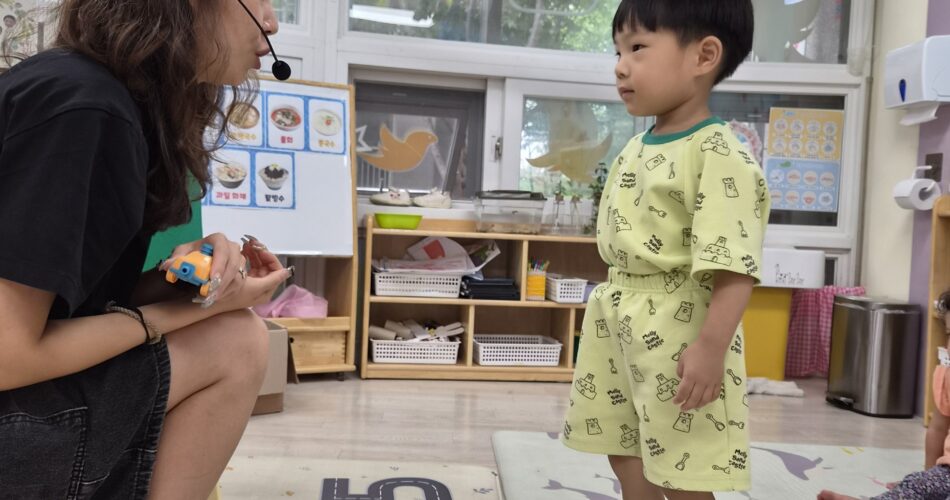 어린이집 교실 안, 연두색 상하복을 입은 어린 지호가 숫자와 도로 그림이 있는 바닥 매트 위에 맨발로 서서, 빨간 머리띠를 한 선생님을 마주보고 있다. 선생님은 지호의 눈높이에 맞춰 무릎을 굽히고 앉아 작은 장난감을 들고 지호와 이야기하고 있다. 지호는 선생님을 집중해서 올려다보고 있다.