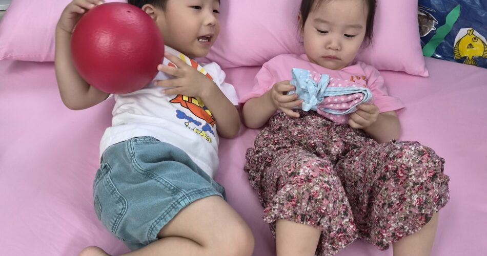 분홍색 침대 시트 위에서 지호와 여자아이가 나란히 누워 놀고 있다. 지호는 빨간 공을 들고 있고, 여자아이는 파란색 물건을 만지고 있다. 두 아이 모두 편안한 표정이다.