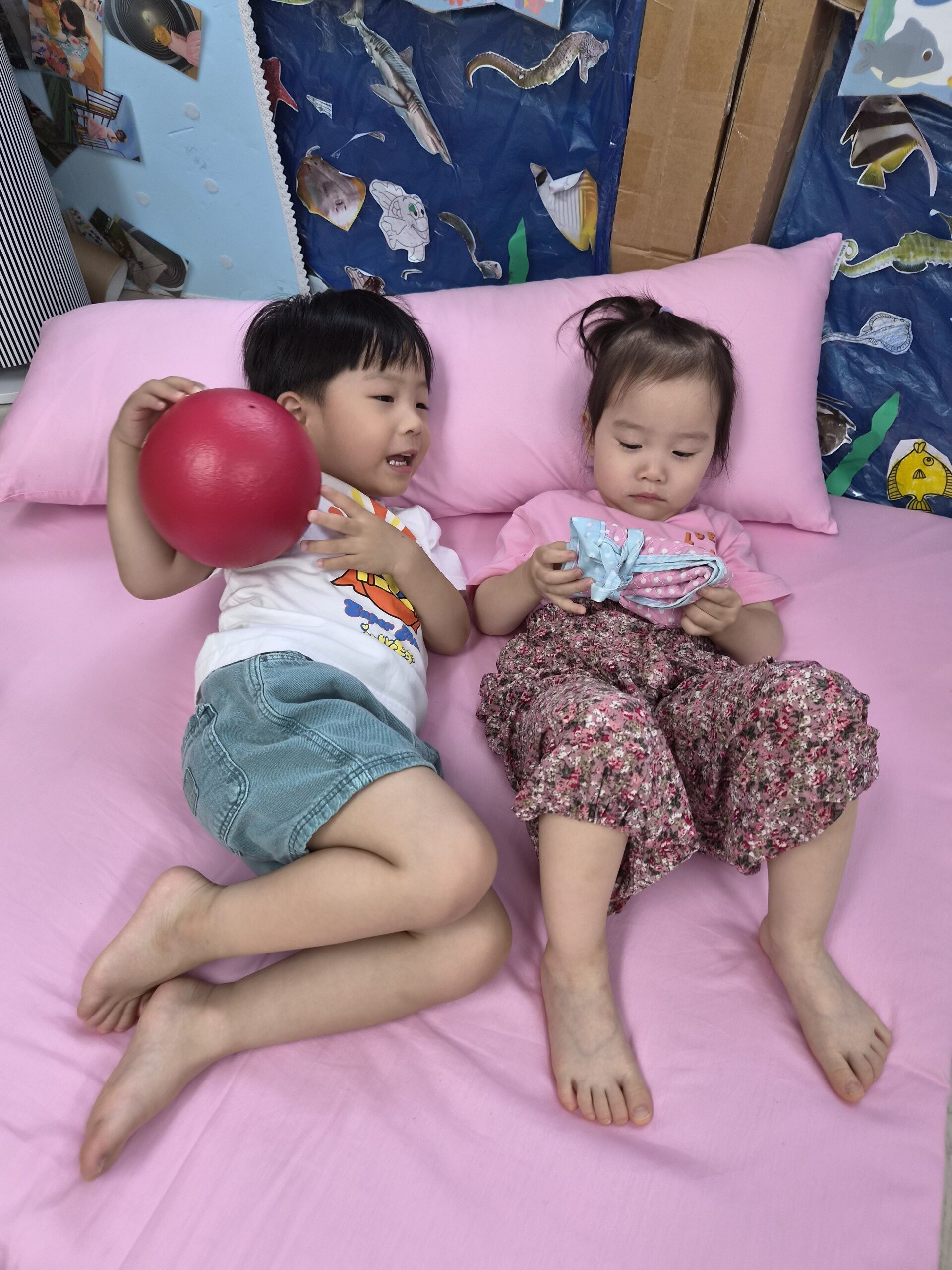 분홍색 침대 시트 위에서 지호와 여자아이가 나란히 누워 놀고 있다. 지호는 빨간 공을 들고 있고, 여자아이는 파란색 물건을 만지고 있다. 두 아이 모두 편안한 표정이다.