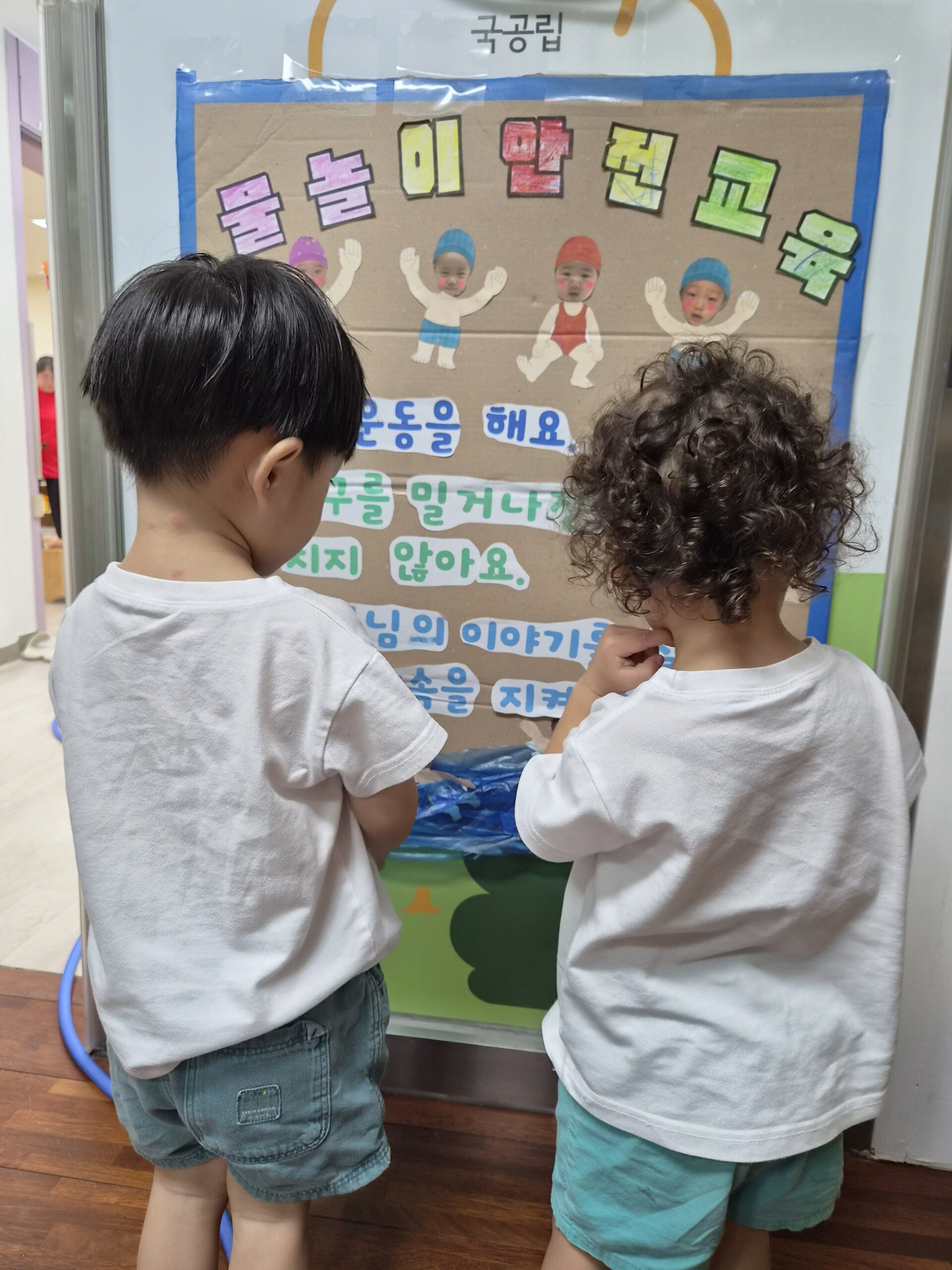 하얀색 티셔츠를 입은 지호가 뒤에서 본 모습으로, 곱슬머리 친구와 함께 '물놀이 안전 교육'이라고 적힌 갈색 종이 게시판을 유심히 바라보고 있는 모습. 게시판에는 귀여운 물놀이 안전 수칙 그림이 그려져 있다.