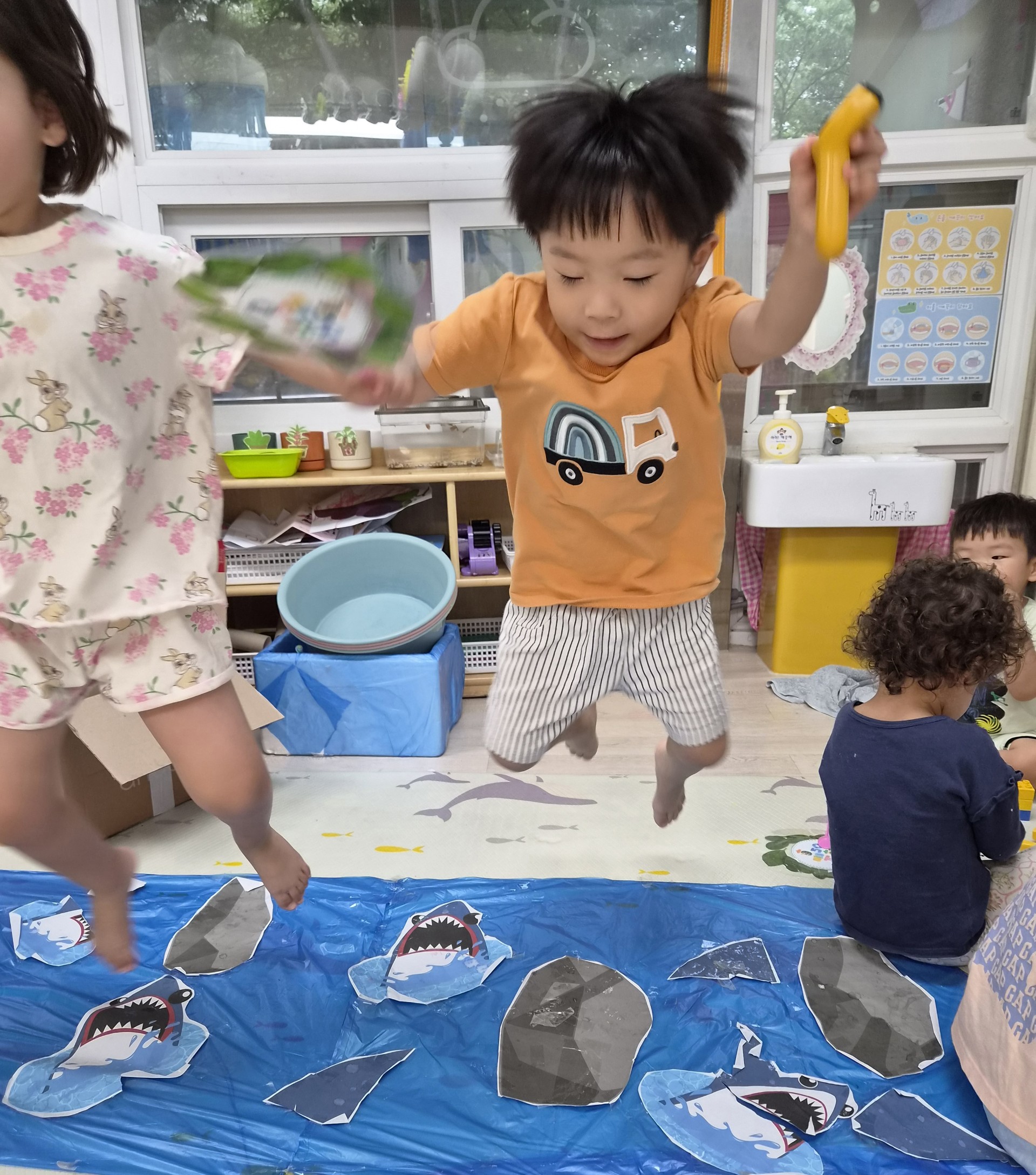 어린이집 실내 바닥에 파란색 천과 상어, 바위 그림이 놓여 있고, 주황색 티셔츠를 입은 지호와 다른 아이가 공중으로 높이 점프하며 즐거워하고 있다. 지호는 노란색 막대 장난감을 들고 있다.