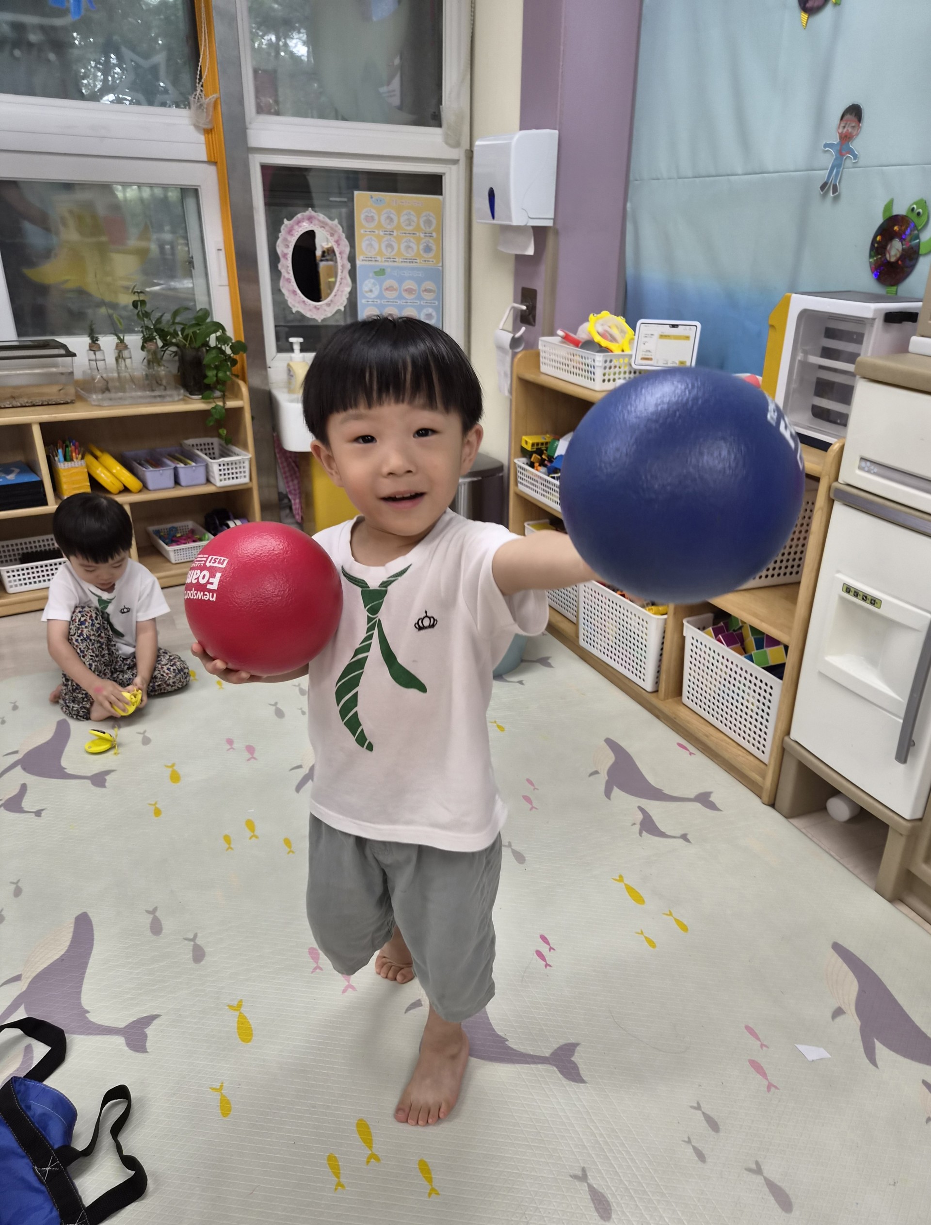 흰색 티셔츠와 회색 반바지를 입은 맨발의 지호가 미소 지으며 카메라를 바라보고 있다. 오른손에는 파란색 공을, 왼손에는 빨간색 공을 들고 있다. 뒤에는 다른 아이가 앉아 있다.