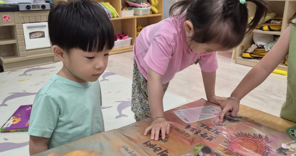 교실에서 지호가 다른 친구들과 함께 테이블에 놓인 다양한 그림과 글자들이 인쇄된 판을 진지하게 들여다보고 있다. 지호는 민트색 티셔츠를 입고 있다.