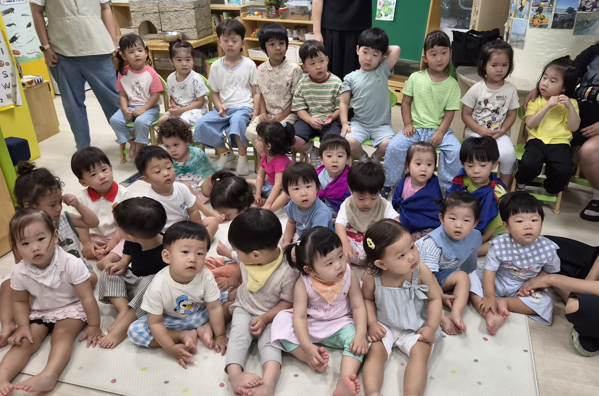 약 20여 명의 어린이집 친구들이 바닥과 작은 의자에 앉아 카메라를 바라보고 있다. 다양한 인종과 표정의 아이들이 한데 모여 있는 모습이 사랑스럽다.