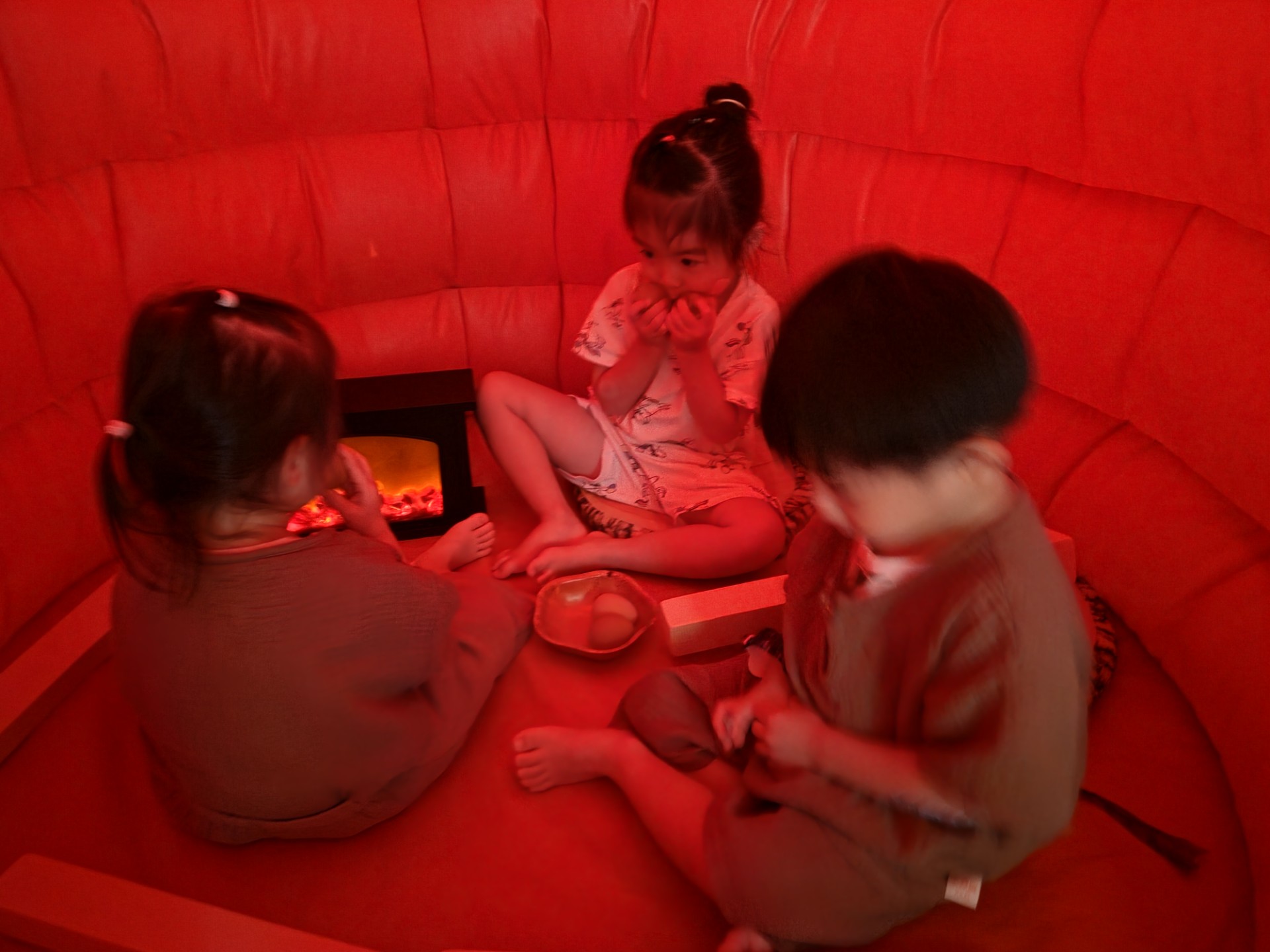 붉은 조명이 비치는 아늑한 공간에 세 명의 아이들이 둥글게 앉아있다. 가운데 지호는 밝은색 옷을 입고 손으로 입을 가린 채 미소 짓고 있으며, 왼쪽 아이는 가상의 벽난로를 바라보고, 오른쪽 아이는 바닥을 응시하며 몰입하고 있다.