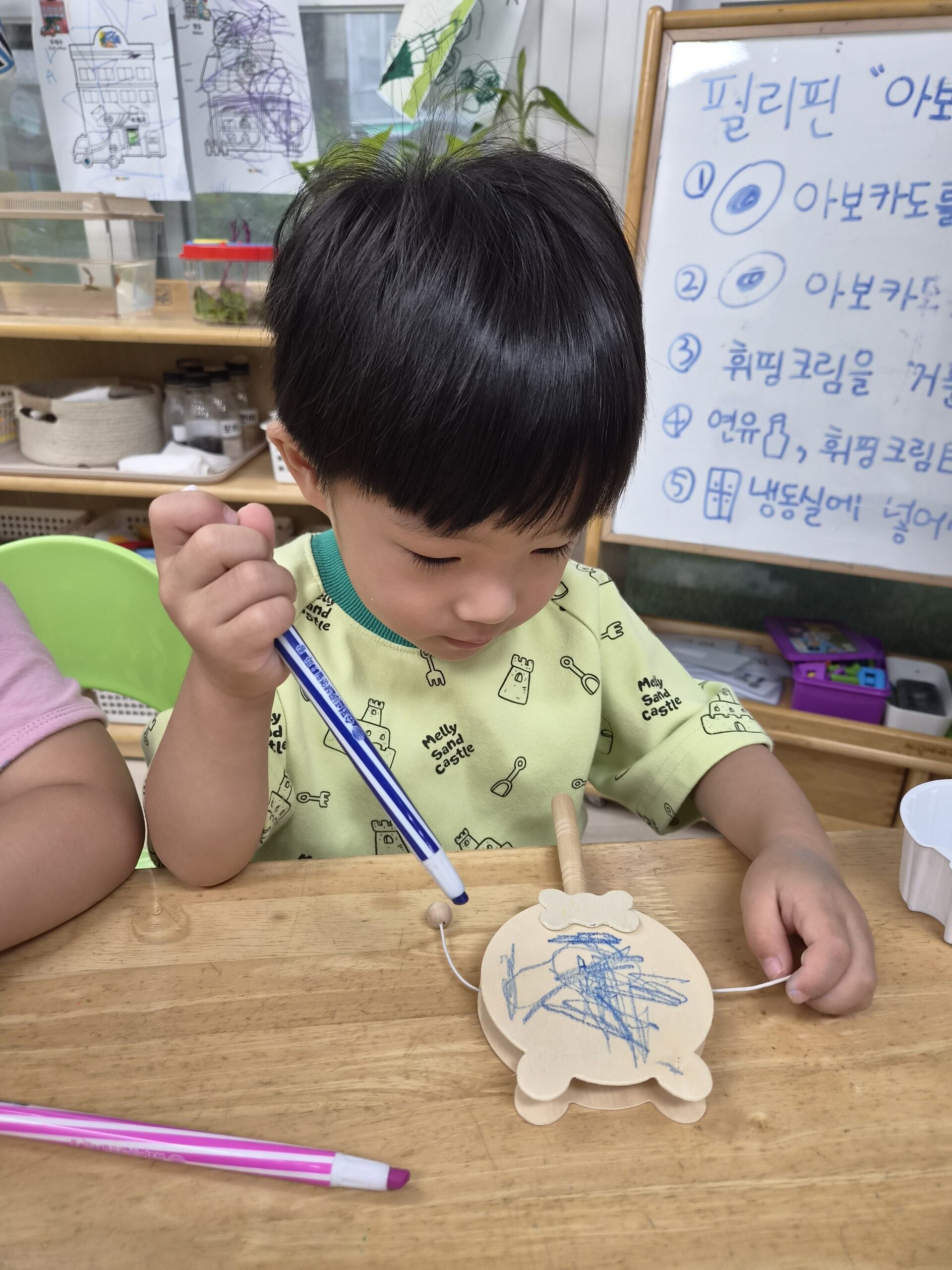 어린이집 교실에서 지호가 나무 탁자에 앉아 연두색 티셔츠를 입고 파란색 마커 펜으로 나무로 만든 거북이 모양 공예품에 그림을 그리는 데 집중하고 있다. 뒤편에는 화이트보드가 보인다.