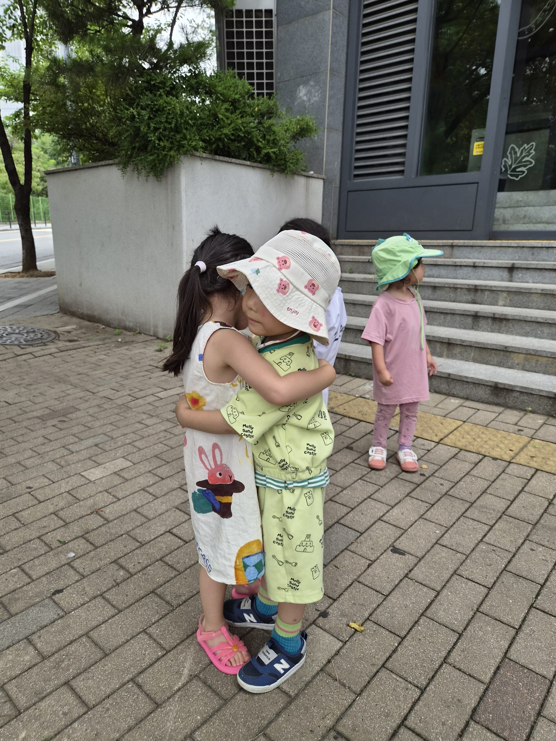 초록색 옷과 체리 무늬 모자를 쓴 지호가 하얀 원피스를 입은 여자아이와 길 위에서 서로 꼭 안고 있다. 다른 아이들이 뒤쪽에 서 있으며, 배경에는 건물과 계단이 보인다.