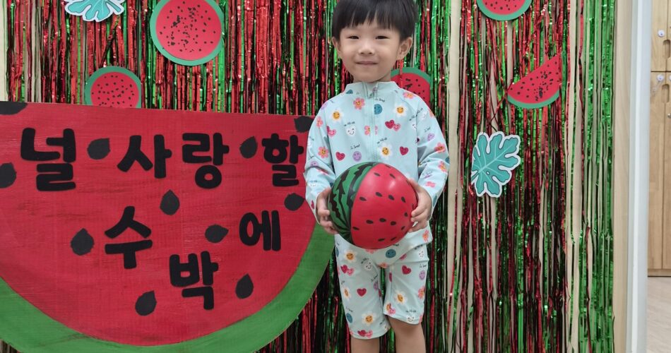 수박 테마 장식 앞에서 연두색 래시가드를 입은 어린 지호가 수박 무늬 공을 들고 서서 카메라를 보고 환하게 웃고 있다. 배경에는 '널 사랑할 수 박'이라고 적힌 커다란 수박 모형과 빨강, 초록색 반짝이 커튼, 수박 조명들이 장식되어 있다.