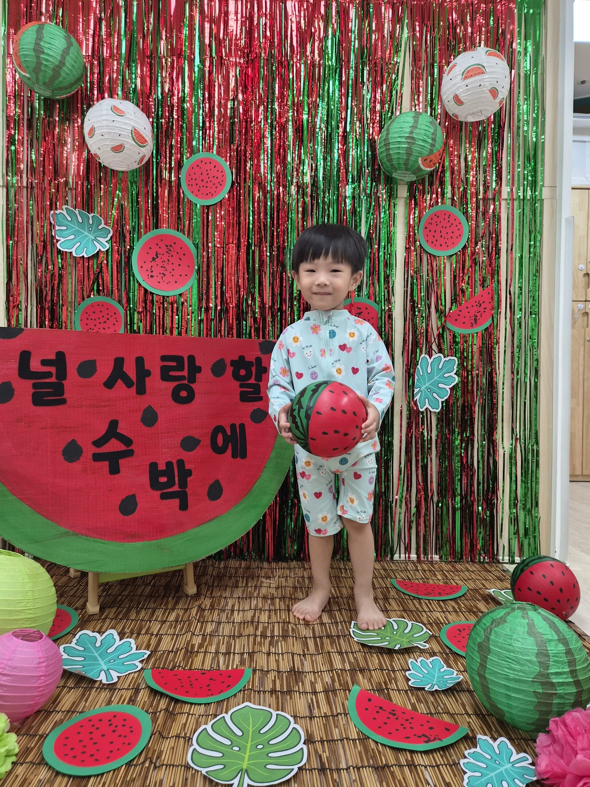 수박 테마 장식 앞에서 연두색 래시가드를 입은 어린 지호가 수박 무늬 공을 들고 서서 카메라를 보고 환하게 웃고 있다. 배경에는 '널 사랑할 수 박'이라고 적힌 커다란 수박 모형과 빨강, 초록색 반짝이 커튼, 수박 조명들이 장식되어 있다.