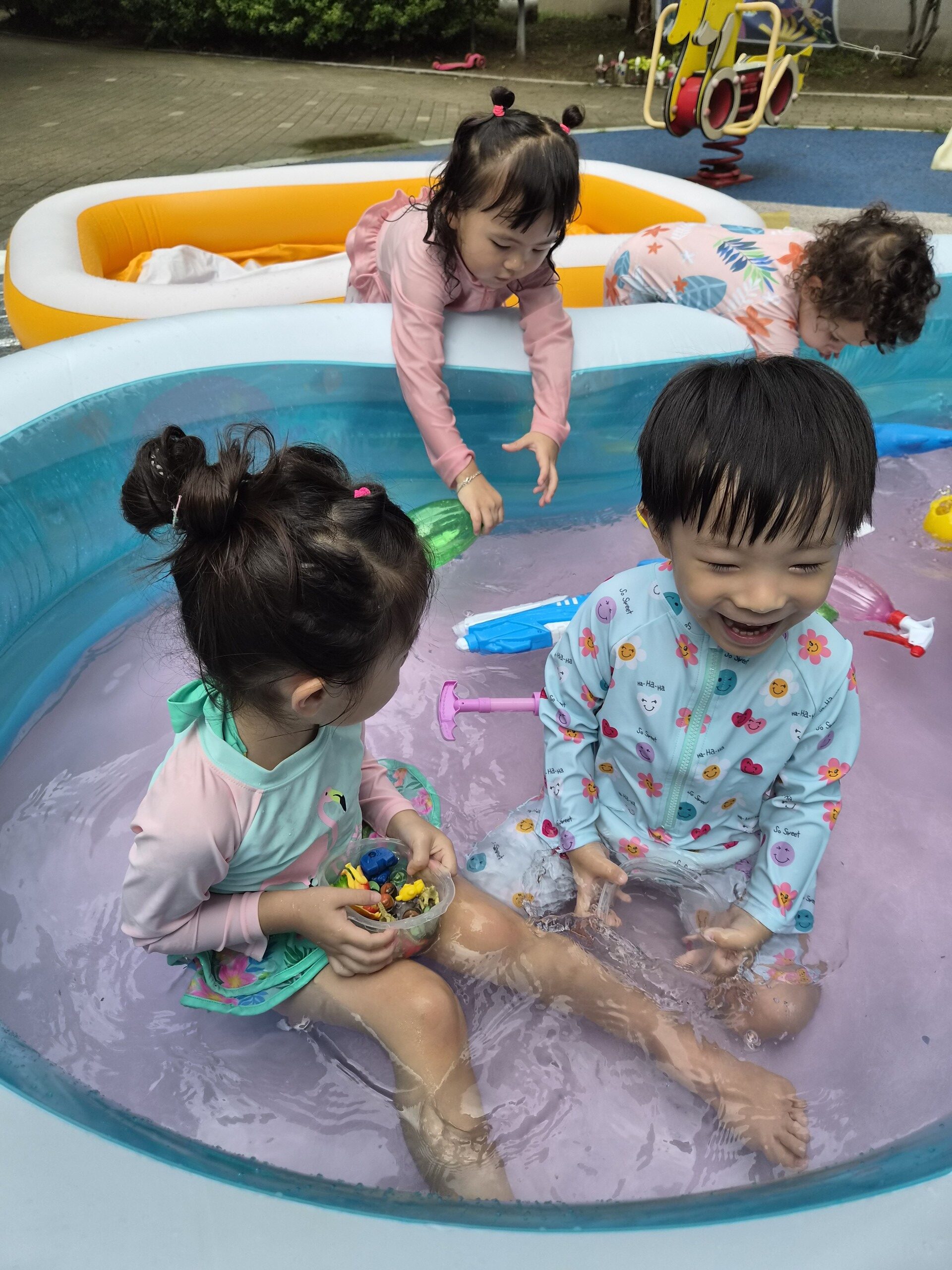분홍색 물이 담긴 수영장 안에 앉아 옆에 앉은 친구와 마주 보며 활짝 웃고 있는 지호. 친구는 작은 장난감들을 들고 있다.
