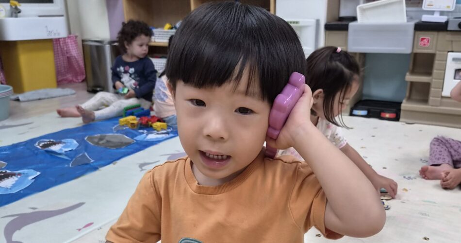 주황색 트럭 그림 티셔츠를 입은 남자아이 지호가 보라색 장난감 전화기를 귀에 대고 활짝 웃으며 정면을 바라보고 있다. 배경에는 다른 아이들과 어린이집 교실의 장난감 부엌이 보인다.