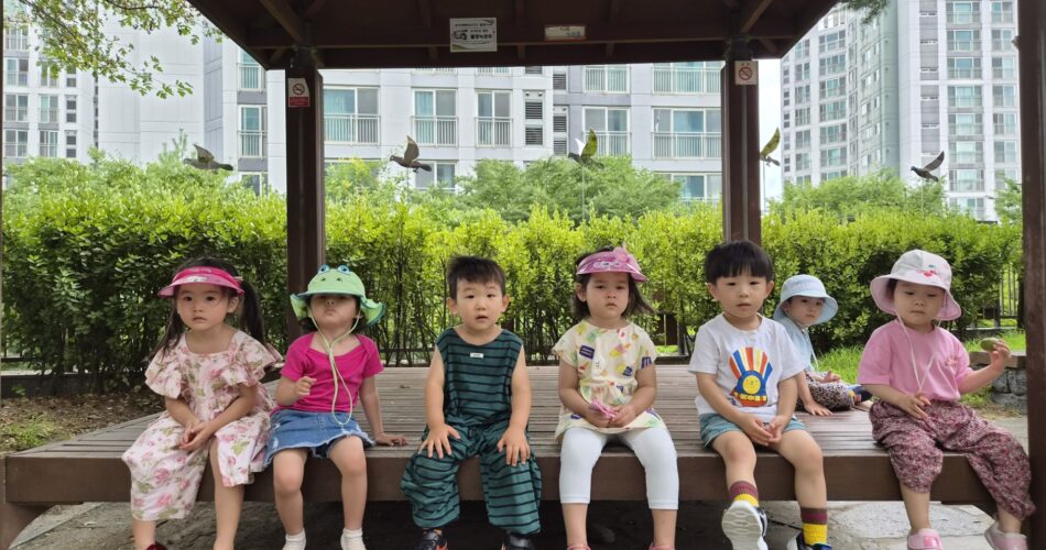 숲과 아파트가 보이는 배경의 정자 아래 벤치에 일곱 명의 어린이집 아이들이 나란히 앉아 카메라를 보고 있다. 모두 모자나 양산을 쓰고 있으며 밝고 편안한 옷차림이다.