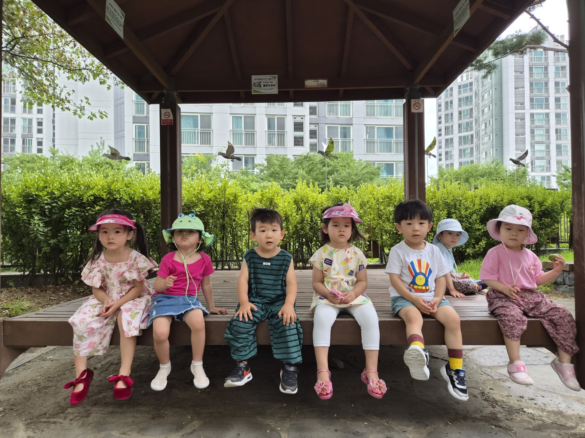 숲과 아파트가 보이는 배경의 정자 아래 벤치에 일곱 명의 어린이집 아이들이 나란히 앉아 카메라를 보고 있다. 모두 모자나 양산을 쓰고 있으며 밝고 편안한 옷차림이다.